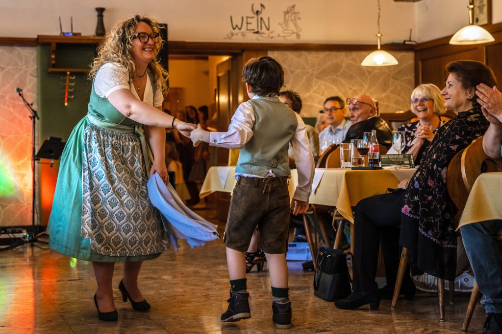Eine Frau und ein Junge in traditioneller Trachtenmode tanzen und lachen gemeinsam bei einer Modenschau in einem gemütlichen Gasthaus. Die Frau trägt ein mintgrünes Dirndl mit gemustertem Rock und hält ein Tuch in der Hand, während der Junge Lederhose, Weste und Hemd trägt. Die Gäste an den Tischen beobachten die Szene aufmerksam und applaudieren, während warmes Licht und die rustikale Einrichtung für eine festliche Atmosphäre sorgen.