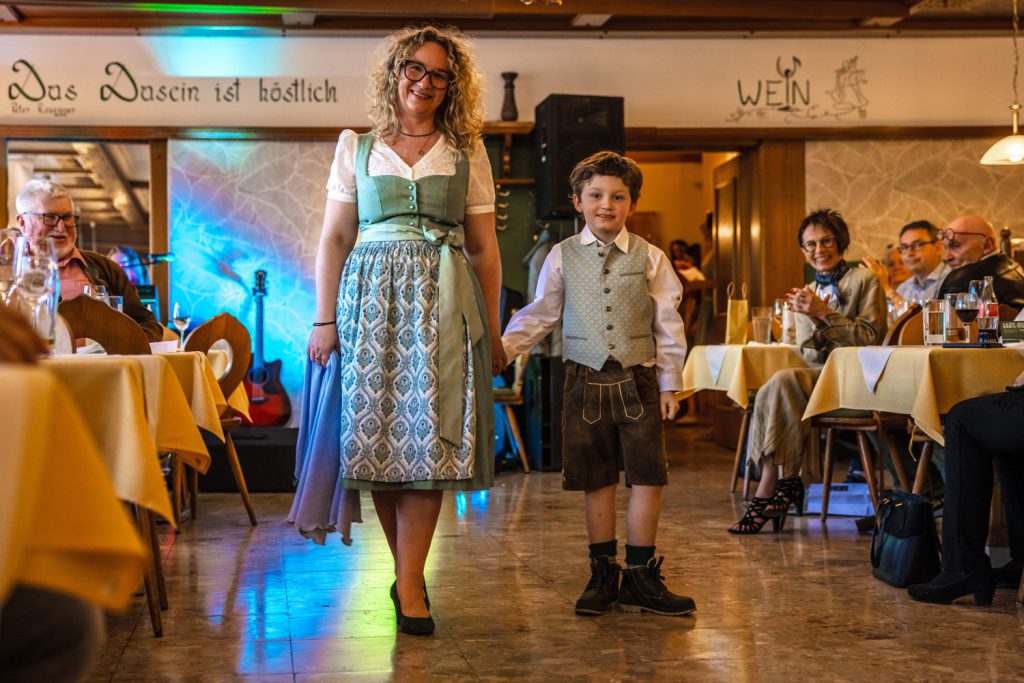 Eine Frau und ein Junge präsentieren gemeinsam traditionelle Trachtenmode bei einer Modenschau in einem gemütlichen Gasthaus. Die Frau trägt ein mintgrünes Dirndl mit gemustertem Rock und hält ein Tuch in der Hand, während der Junge in Lederhose, Hemd und Weste neben ihr läuft. Die Gäste sitzen an den Tischen und verfolgen die Modenschau aufmerksam, während bunte Lichter und eine Gitarre im Hintergrund für eine festliche Stimmung sorgen.