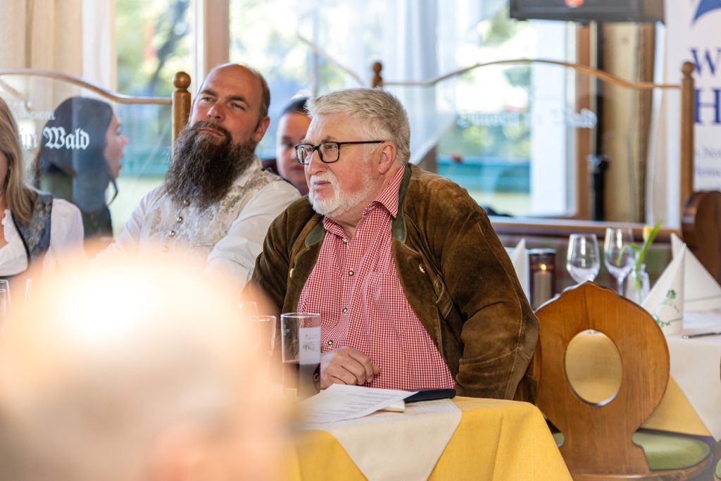 Zwei Männer sitzen bei einer Veranstaltung an einem gedeckten Tisch und hören aufmerksam zu. Im Vordergrund sitzt ein älterer Mann mit Brille, rotem karierten Hemd und brauner Jacke, daneben ein Mann mit langem Bart und traditionell wirkendem Hemd. Die Stimmung wirkt ruhig und konzentriert.