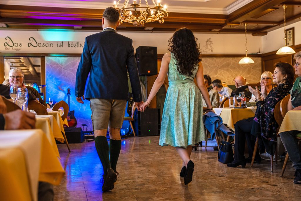 Ein junges Paar präsentiert Trachtenmode bei einer Modenschau in einem traditionellen Gasthaus und geht Hand in Hand über den Laufsteg. Der Mann trägt ein dunkelblaues Sakko mit heller Lederhose und grünen Kniestrümpfen, die Frau ein mintgrünes Kleid und hält eine blaue Jacke in der Hand. Die Gäste sitzen an den gedeckten Tischen, applaudieren und verfolgen die Modenschau in festlicher Atmosphäre mit bunten Lichteffekten im Hintergrund.