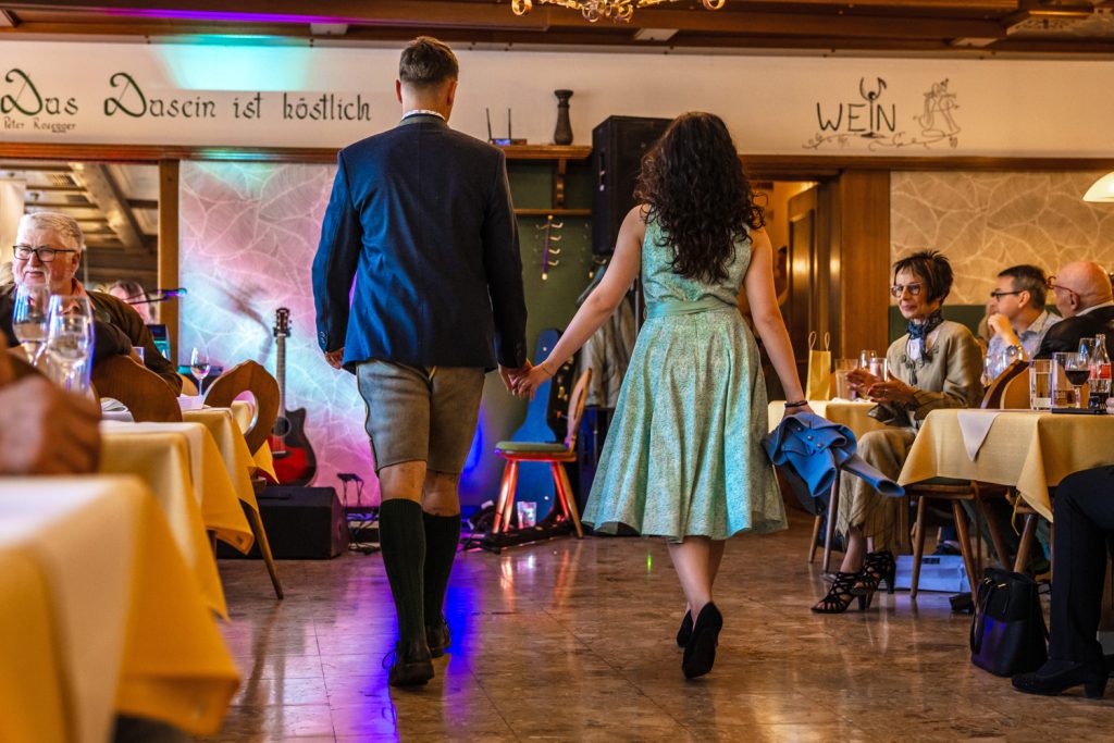 Ein junges Paar präsentiert Trachtenmode bei einer Modenschau in einem traditionellen Gasthaus und geht Hand in Hand über den Laufsteg. Der Mann trägt ein dunkelblaues Sakko mit Lederhose und grünen Kniestrümpfen, die Frau ein mintgrünes Kleid mit passender Schleife und hält eine blaue Jacke in der Hand. Die Gäste sitzen an gedeckten Tischen und verfolgen die Modenschau in stimmungsvoller Atmosphäre mit bunten Lichteffekten im Hintergrund.