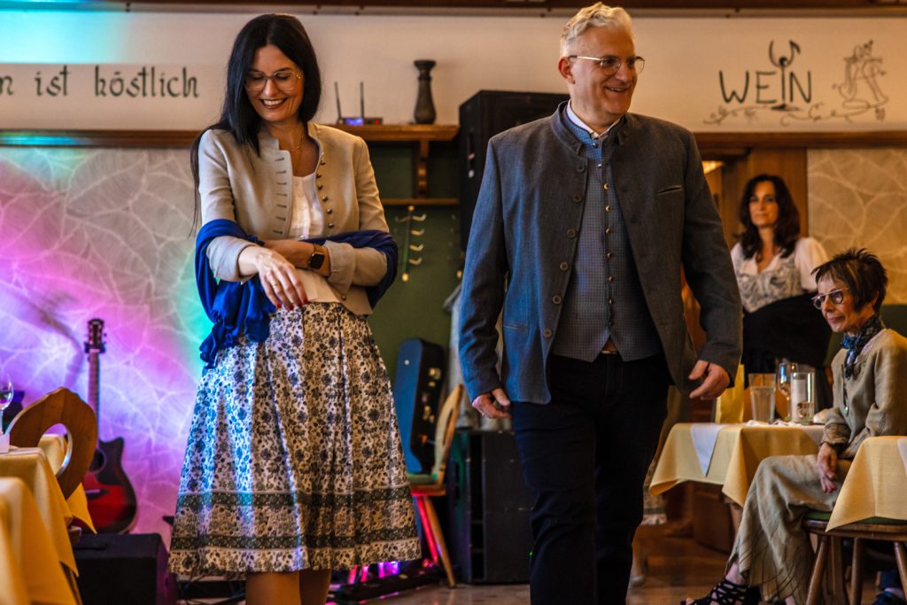 Ein Paar präsentiert moderne Trachtenmode bei einer Modenschau in einem traditionellen Gasthaus. Die Frau trägt einen gemusterten Rock mit heller Bluse, kurzem Blazer und blauem Schal, während der Mann ein elegantes Trachtensakko mit passender Weste und dunkler Hose kombiniert. Beide lächeln während ihres Laufstegs, begleitet von stimmungsvoller Beleuchtung und aufmerksam zuschauenden Gästen an den Tischen.