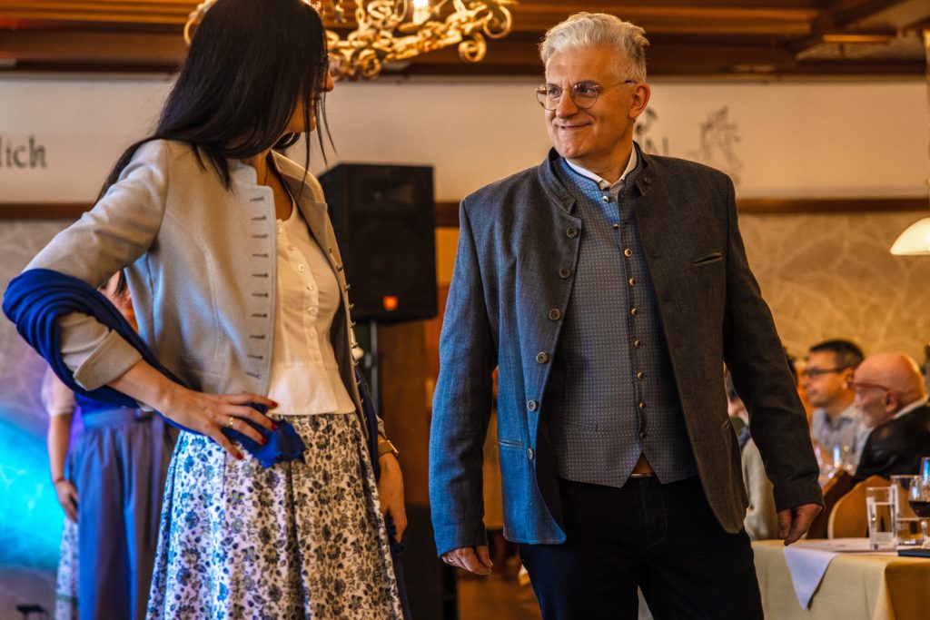 Ein Paar präsentiert elegante Trachtenmode bei einer Modenschau in einem traditionellen Gasthaus. Die Frau trägt einen gemusterten Rock mit heller Bluse, kurzem Blazer und blauem Schal, während der Mann ein klassisches Trachtensakko mit passender Weste und dunkler Hose kombiniert. Beide lächeln sich an, während die Gäste an den gedeckten Tischen die Vorführung aufmerksam verfolgen.