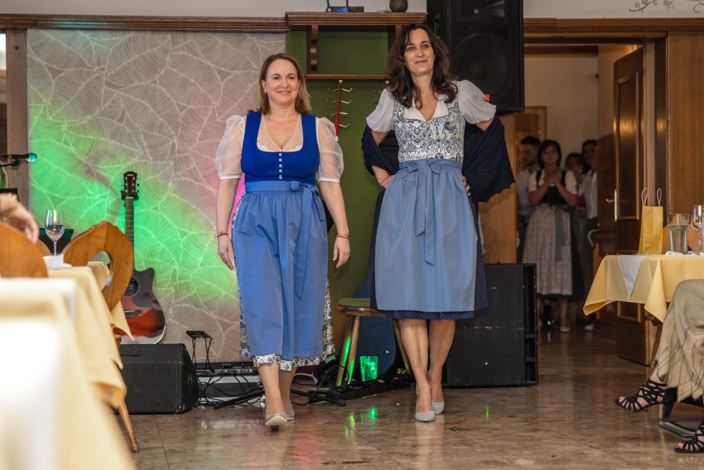 Zwei Frauen präsentieren stilvolle Dirndl bei einer Trachtenmodenschau in einem traditionellen Gasthaus. Eine trägt ein kräftig blaues Dirndl mit transparenter Puffbluse, die andere ein blau-weiß gemustertes Dirndl mit dunklem Schultertuch und heller Schürze. Im Hintergrund sorgen farbige Lichteffekte, eine Gitarre und die aufmerksam zuschauenden Gäste für eine festliche und gemütliche Atmosphäre.