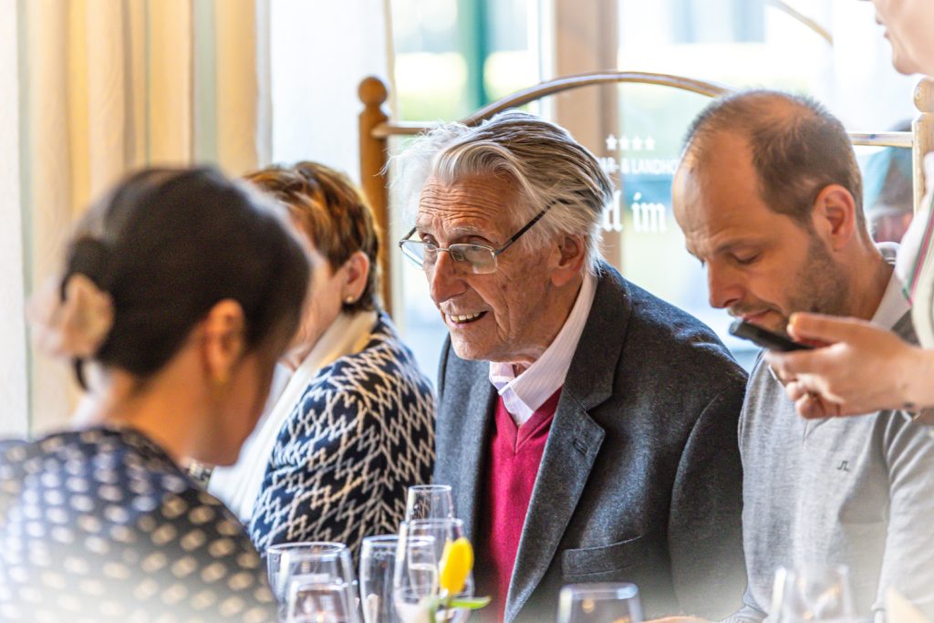 Mehrere Gäste sitzen gemeinsam an einem Tisch in einem hellen Restaurantbereich. Im Mittelpunkt lächelt ein älterer Mann mit grauen Haaren und Brille in die Runde, während andere Gäste aufmerksam zuhören oder auf ihr Smartphone blicken. Die Atmosphäre wirkt freundlich und gesellig.