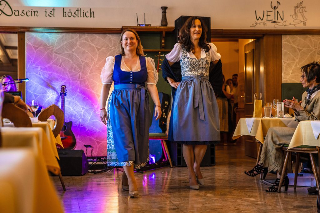 Zwei Frauen präsentieren elegante Dirndl bei einer Trachtenmodenschau in einem gemütlichen Gasthaus. Eine trägt ein kräftig blaues Dirndl mit transparenter Bluse und gemusterter Schürze, die andere ein helles Dirndl mit blau-weißem Muster und dunklem Schultertuch. Im Hintergrund sorgen bunte Lichteffekte und eine Gitarre für eine stimmungsvolle Atmosphäre, während die Gäste an den Tischen aufmerksam zusehen.
