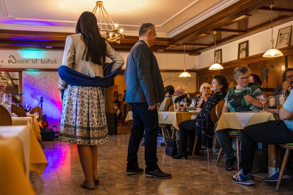Ein Paar präsentiert bei einer Trachtenmodenschau elegante traditionelle Mode und läuft gemeinsam durch einen gemütlichen Gastraum. Die Frau trägt einen gemusterten Rock mit hellem Blazer und blauem Schal, der Mann ein klassisches Trachtensakko mit Weste und dunkler Hose. Die Gäste sitzen an gedeckten Tischen und verfolgen aufmerksam die Modenschau, während farbige Beleuchtung und rustikale Holzelemente für eine festliche Stimmung sorgen.