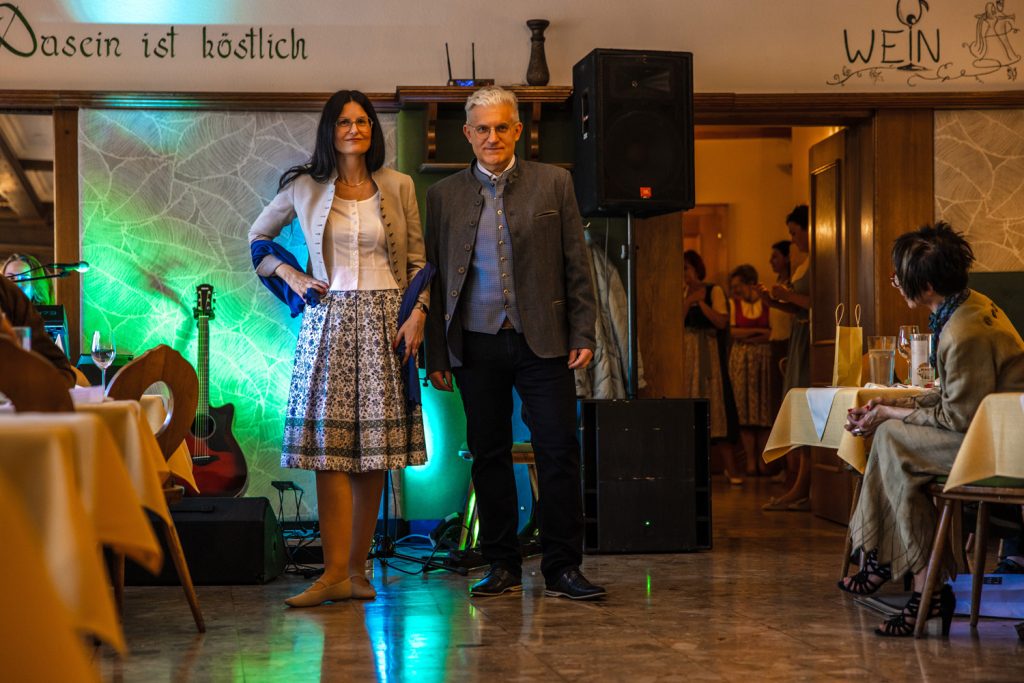 Ein Mann und eine Frau stehen bei einer Trachtenmodenschau gemeinsam auf dem Laufsteg eines gemütlichen Gastraums. Die Frau trägt ein traditionelles Outfit mit gemustertem Rock, heller Jacke und Schal, der Mann ein elegantes Trachtensakko mit Weste. Farbige Beleuchtung, eine Gitarre im Hintergrund und die aufmerksam zuschauenden Gäste verleihen der Veranstaltung eine festliche Stimmung.