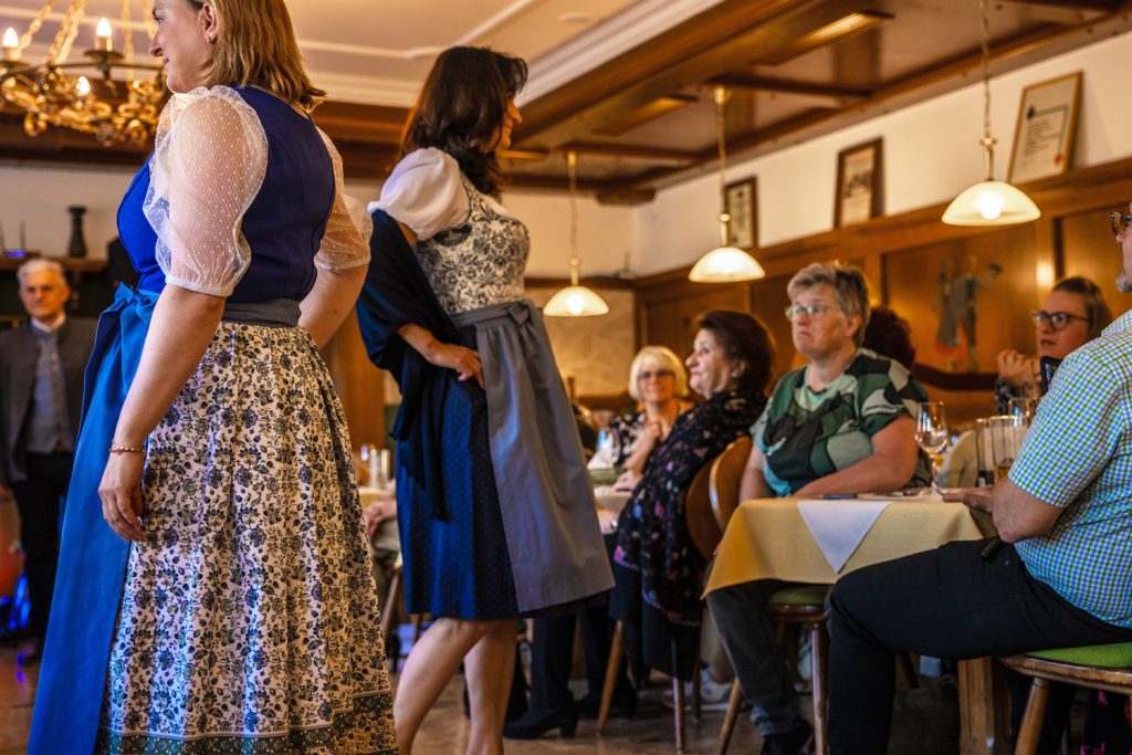 Zwei Frauen präsentieren bei einer Trachtenmodenschau stilvolle Dirndl in Blau- und Grautönen. Fotografiert seitlich zeigen die Kleider feine Muster, transparente Ärmel und elegante Schürzen. Die Gäste sitzen aufmerksam im gemütlichen Gastraum mit Holzvertäfelung und warmem Licht und verfolgen die Modenschau interessiert.