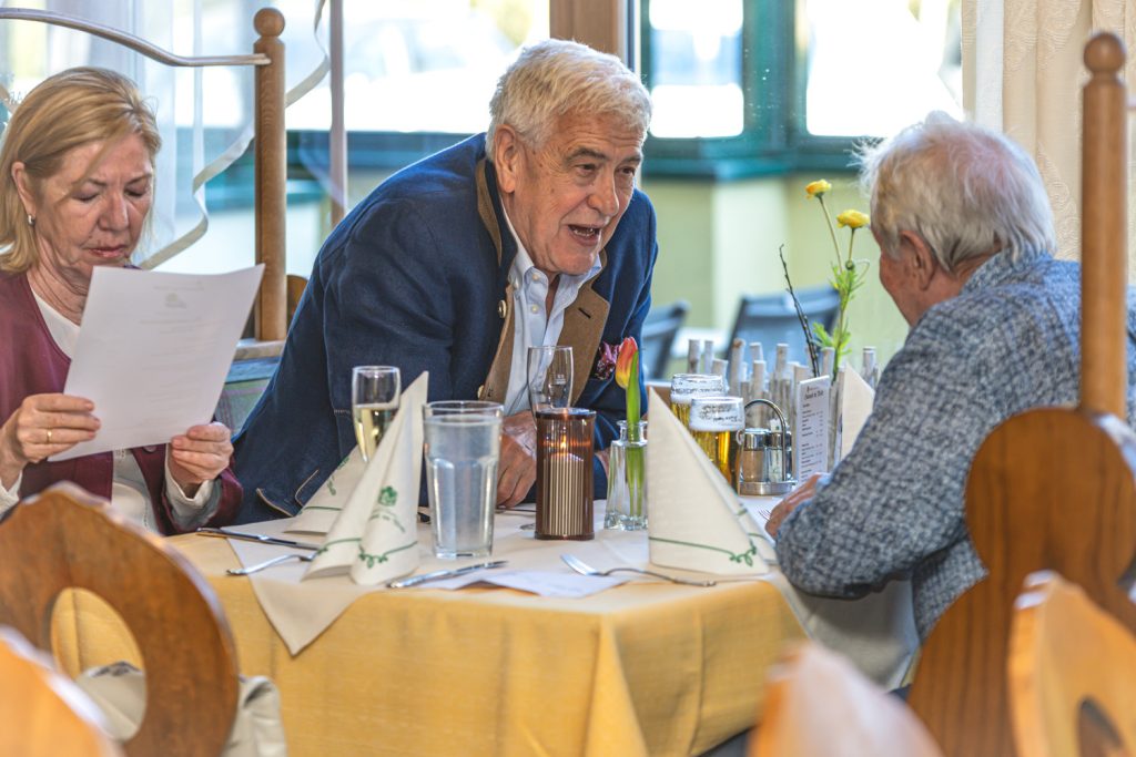 Mehrere Gäste sitzen an einem festlich gedeckten Tisch in einem Restaurant und unterhalten sich angeregt. Ein älterer Mann in blauem Sakko spricht lebhaft mit einem anderen Gast, während eine Frau daneben aufmerksam ein Blatt Papier liest. Auf dem Tisch stehen Getränke, Servietten und kleine Blumendekorationen, was eine gemütliche und gesellige Atmosphäre vermittelt.