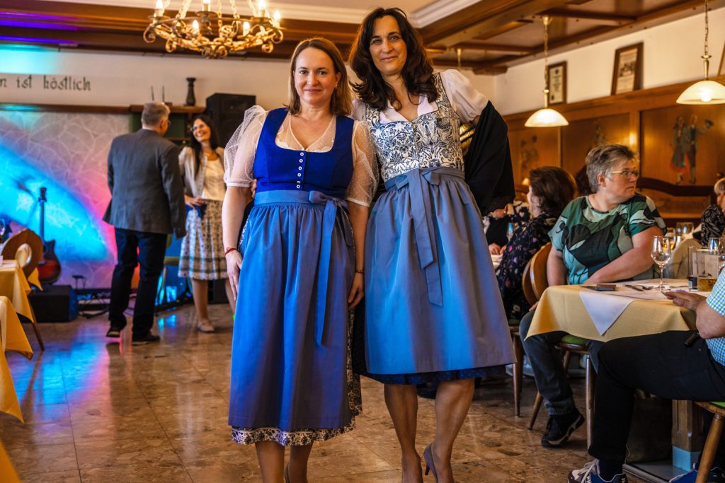Zwei Frauen präsentieren bei einer Trachtenmodenschau elegante Dirndl in kräftigen Blau- und Grautönen. Sie stehen lächelnd nebeneinander im Mittelpunkt des Laufstegs, während die Gäste an den Tischen die traditionelle Mode aufmerksam betrachten. Der gemütliche Gastraum mit Holzelementen, Kronleuchtern und farbiger Beleuchtung sorgt für eine festliche Atmosphäre.