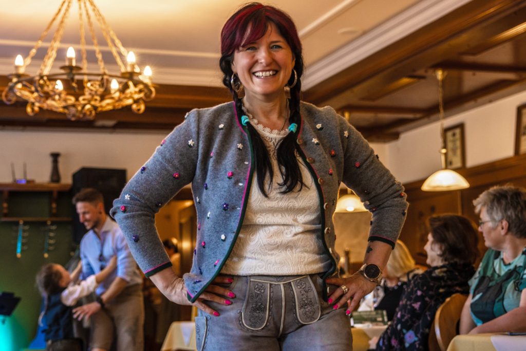 Eine lachende Frau präsentiert bei einer Trachtenmodenschau ein modernes Outfit mit grauer, bestickter Strickjacke, heller Spitzenbluse und kurzer Lederhose. Sie posiert selbstbewusst mit den Händen in der Hüfte in einem gemütlichen Gastraum mit Kronleuchtern und Holzvertäfelung. Im Hintergrund sitzen Gäste an den Tischen und beobachten die Veranstaltung aufmerksam.
