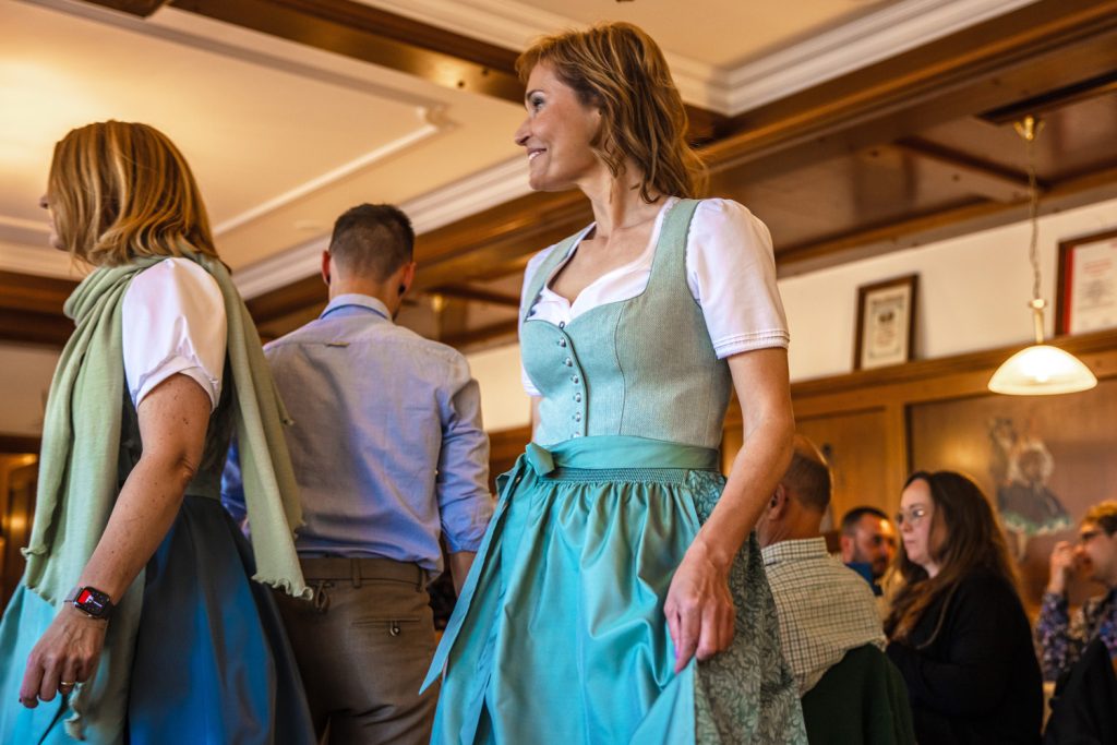 Eine Frau präsentiert bei einer Trachtenmodenschau ein elegantes Dirndl in Türkis- und Mintgrün mit weißer Bluse und passender Schleife. Neben ihr läuft eine weitere Frau im Dirndl vorbei, während Gäste im gemütlichen Gastraum aufmerksam zuschauen. Die warme Beleuchtung und die Holzvertäfelung verleihen der Szene eine festliche und traditionelle Atmosphäre.