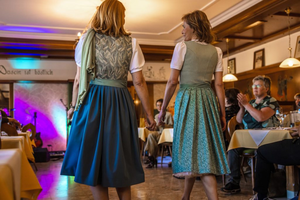 Zwei Frauen laufen Hand in Hand über den Laufsteg einer Trachtenmodenschau und präsentieren Dirndl in harmonischen Blau- und Grüntönen. Fotografiert von hinten zeigt das Bild die feinen Stoffdetails und Schleifen der Kleider. Im stimmungsvoll beleuchteten Gastraum verfolgen die Gäste aufmerksam die Modenschau und applaudieren begeistert.