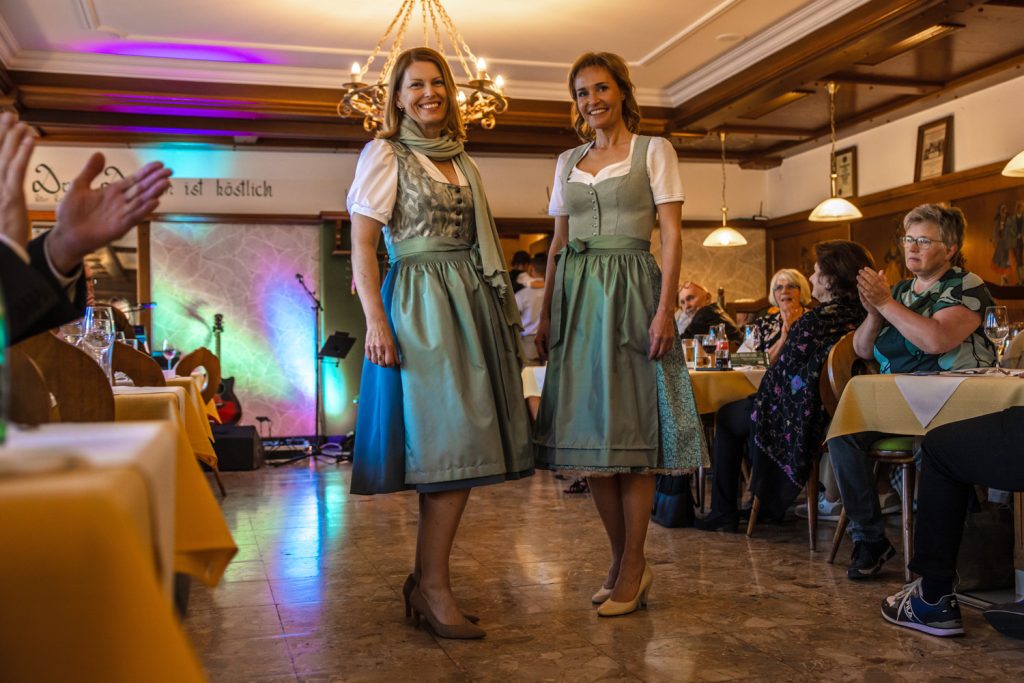 Zwei Frauen präsentieren bei einer Trachtenmodenschau elegante Dirndl in sanften Grün- und Türkistönen. Sie stehen lächelnd in einem gemütlichen Gastraum mit Holzelementen und Kronleuchtern, während die Gäste an den Tischen applaudieren. Im Hintergrund sorgen bunte Lichter und eine kleine Bühne für festliche Stimmung.