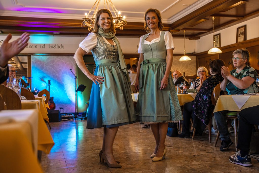Zwei Frauen in türkisfarbenen Dirndln stehen bei einer Veranstaltung in einem Gastraum und präsentieren ihre Trachtenoutfits. Eine Frau trägt zusätzlich einen Schal. Gäste sitzen an den Tischen und applaudieren. Im Hintergrund sind bunte Bühnenbeleuchtung und eine Gitarre zu sehen.