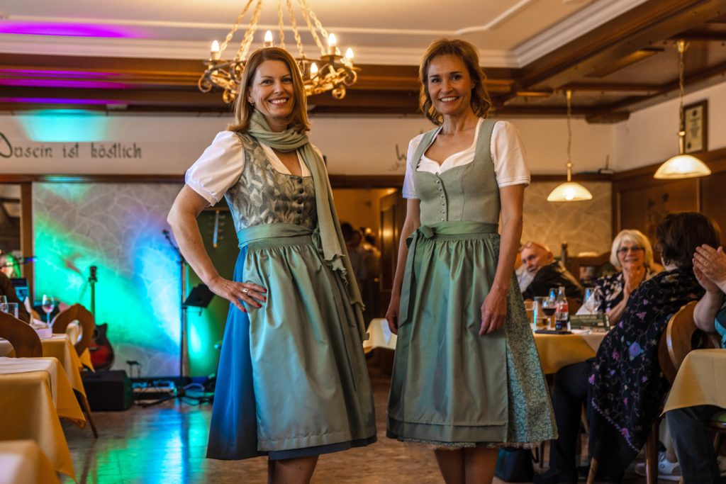 Zwei Frauen in türkisfarbenen Dirndln stehen bei einer Veranstaltung in einem Gastraum und präsentieren ihre Trachtenoutfits. Eine Frau trägt zusätzlich einen passenden Schal. Gäste sitzen an den Tischen und beobachten die Szene. Im Hintergrund sind bunte Bühnenbeleuchtung und eine Gitarre zu sehen.