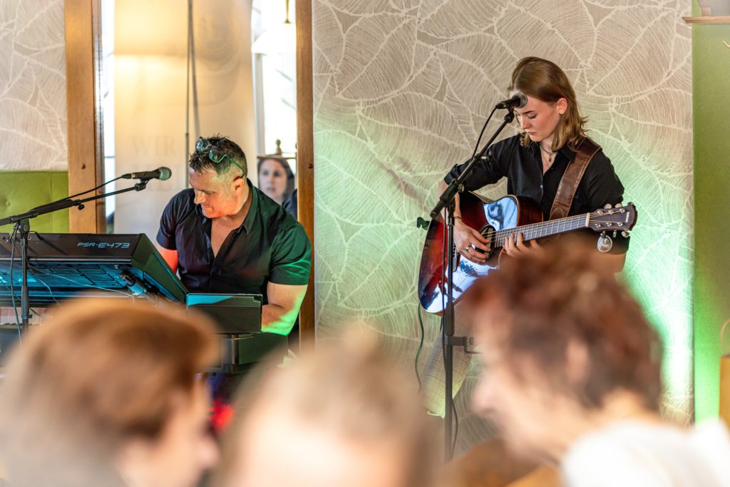 Zwei Musiker treten bei einer Veranstaltung auf: Ein Mann spielt Keyboard, während eine junge Frau daneben akustische Gitarre spielt. Beide stehen beziehungsweise sitzen vor Mikrofonen, umgeben von stimmungsvoller Beleuchtung. Im Vordergrund sind unscharf die Köpfe der Gäste zu sehen, die der Live-Musik aufmerksam zuhören.