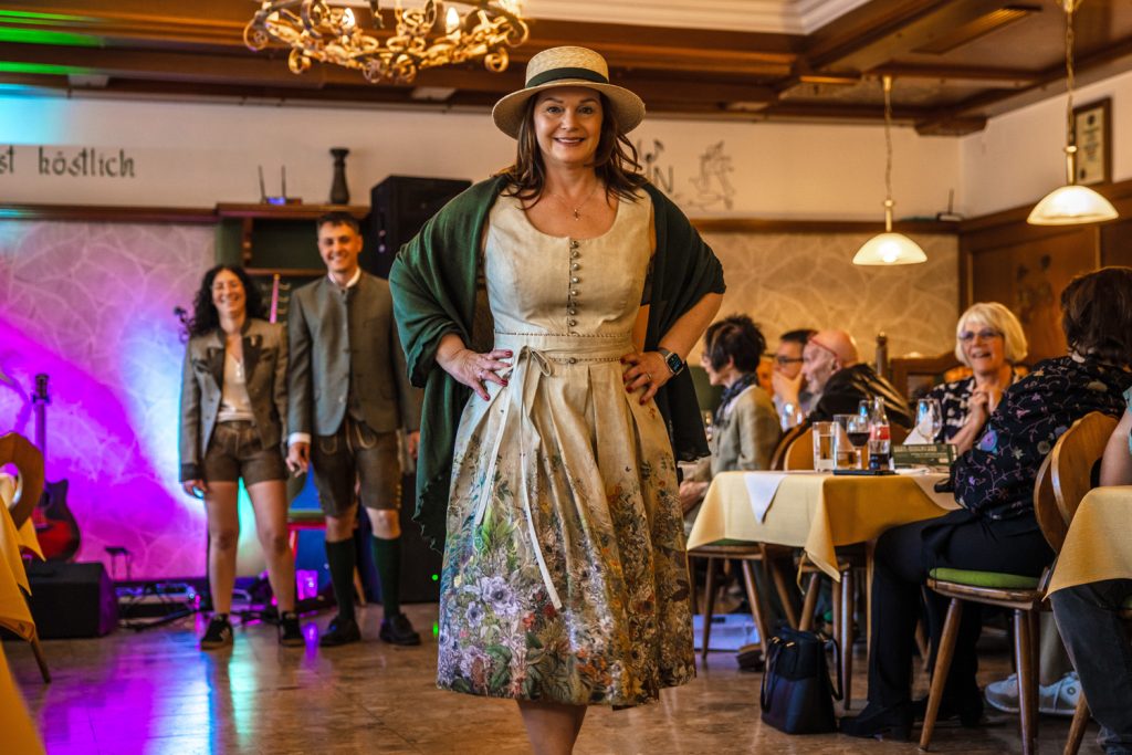 Eine Frau mit Hut und gemustertem Kleid präsentiert bei einer Veranstaltung ein Trachtenoutfit in einem Gastraum. Über den Schultern trägt sie ein dunkles Tuch. Im Hintergrund stehen weitere Personen in Tracht, Gäste sitzen an den Tischen und beobachten die Szene. Der Raum ist mit Holzvertäfelungen und bunter Beleuchtung gestaltet.