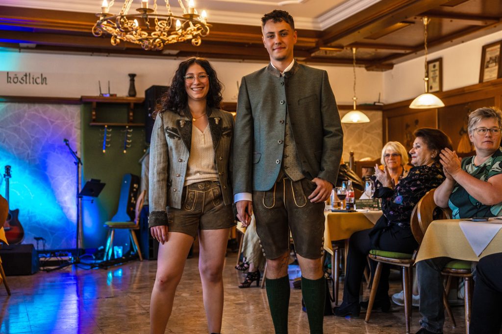 Ein junges Paar in Tracht steht Hand in Hand bei einer Veranstaltung in einem Gastraum und lächelt in die Kamera. Beide tragen Lederhosen, der Mann zusätzlich eine grüne Trachtenjacke, die Frau eine kurze Trachtenjacke und helle Bluse. Gäste sitzen an den Tischen und applaudieren. Im Hintergrund sind eine Bühne mit Beleuchtung und eine Gitarre zu sehen.