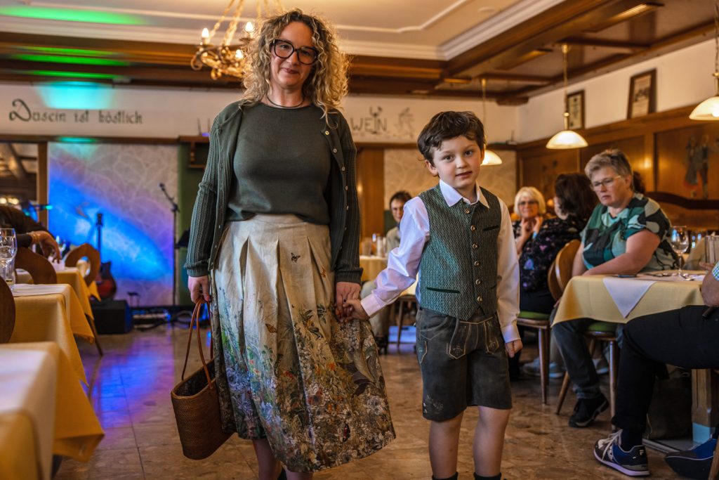 Eine Frau mit lockigen Haaren und ein Junge in Tracht gehen Hand in Hand bei einer Veranstaltung durch einen Gastraum. Die Frau trägt einen langen gemusterten Rock und hält eine Tasche, der Junge trägt Lederhose und Weste. Gäste sitzen an den Tischen und beobachten die Szene. Im Hintergrund sind bunte Bühnenbeleuchtung und eine Gitarre zu sehen.