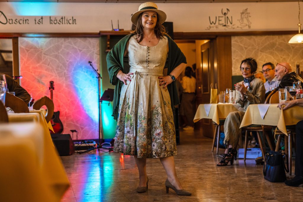 Eine Frau mit Hut und gemustertem Kleid steht bei einer Veranstaltung in einem Gastraum und präsentiert ein Trachtenoutfit. Über den Schultern trägt sie ein dunkles Tuch. Gäste sitzen an den Tischen und beobachten die Szene. Im Hintergrund sind bunte Bühnenbeleuchtung und eine Gitarre zu sehen.