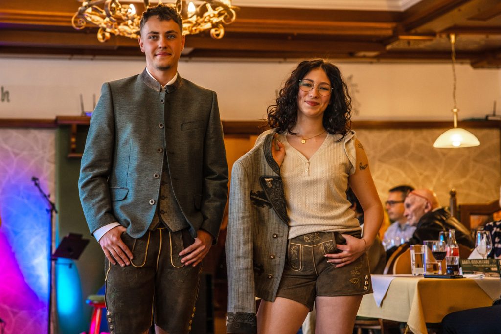 Ein junges Paar in Tracht steht bei einer Veranstaltung in einem Gastraum und präsentiert traditionelle Kleidung. Der Mann trägt Lederhose und eine graue Trachtenjacke, die Frau Lederhose mit heller Bluse und hält eine Jacke über der Schulter. Gäste sitzen an den Tischen und beobachten die Szene. Im Hintergrund ist bunte Bühnenbeleuchtung zu sehen.