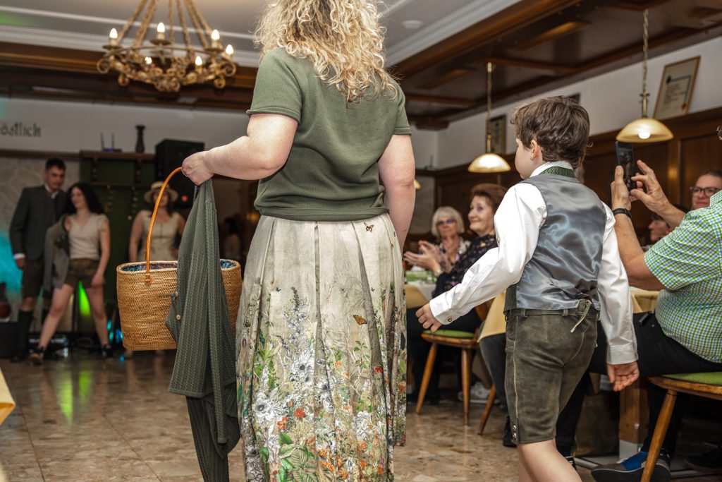 Eine Frau mit lockigen Haaren und ein Junge in Tracht gehen bei einer Veranstaltung durch einen Gastraum. Die Frau trägt einen langen gemusterten Rock und hält eine Tasche sowie eine Jacke in der Hand, der Junge trägt Lederhose und Weste. Gäste sitzen an den Tischen und applaudieren, eine Person fotografiert die Szene mit dem Smartphone.