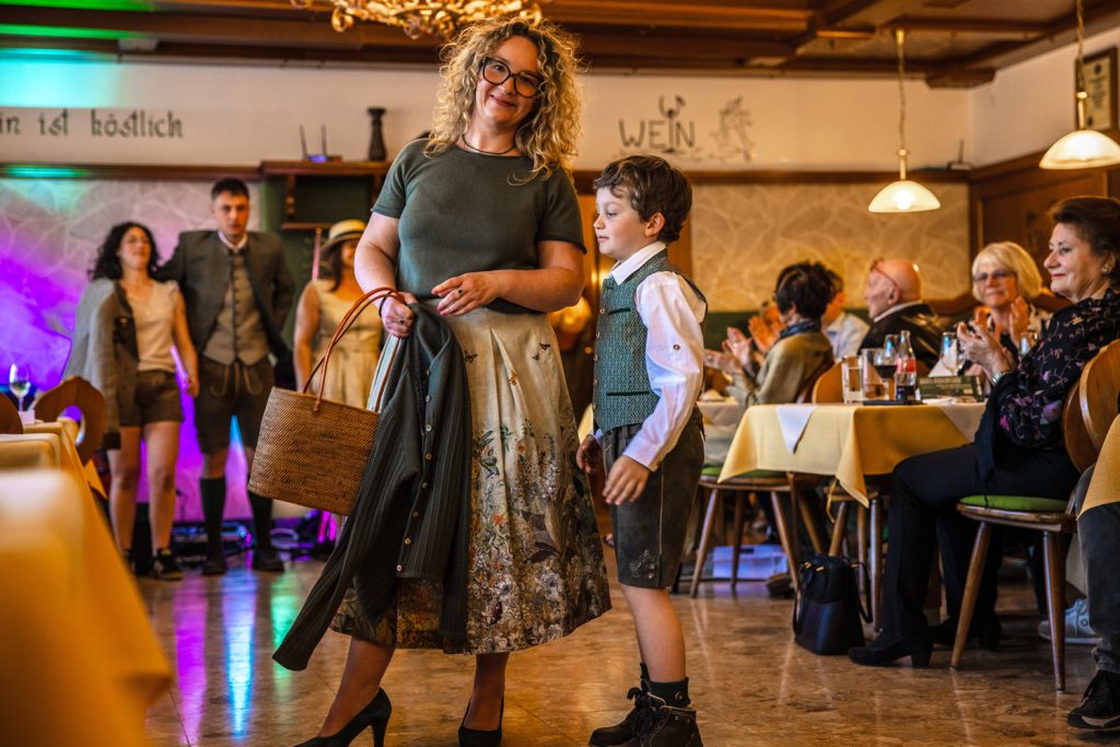 Eine Frau mit lockigen Haaren und ein Junge in Tracht stehen bei einer Veranstaltung in einem Gastraum. Die Frau hält eine Jacke und eine Tasche in der Hand, der Junge trägt Lederhose und Weste. Gäste sitzen an den Tischen und applaudieren. Im Hintergrund sind weitere Personen in Tracht und bunte Bühnenbeleuchtung zu sehen.