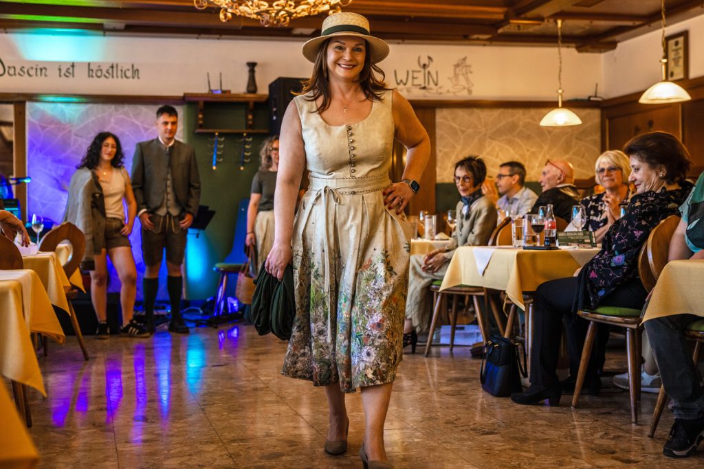 Eine Frau mit Hut und gemustertem Kleid geht lächelnd bei einer Veranstaltung durch einen Gastraum und präsentiert ein Trachtenoutfit. Gäste sitzen an den Tischen und beobachten die Szene. Im Hintergrund stehen weitere Personen in Tracht, der Raum ist mit Holzvertäfelungen und bunter Beleuchtung gestaltet.