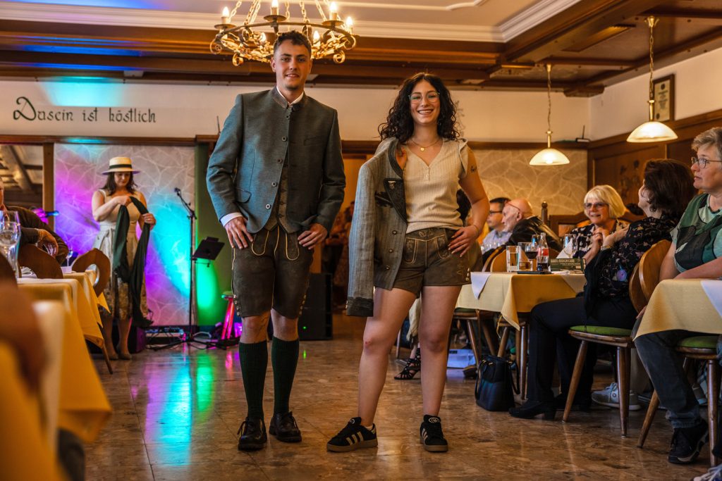 Ein junges Paar in Tracht steht bei einer Veranstaltung in einem Gastraum und präsentiert traditionelle Kleidung. Beide tragen Lederhosen, der Mann zusätzlich eine graue Trachtenjacke. Gäste sitzen an den Tischen und beobachten die Szene. Im Hintergrund sind bunte Bühnenbeleuchtung und weitere Personen zu sehen.