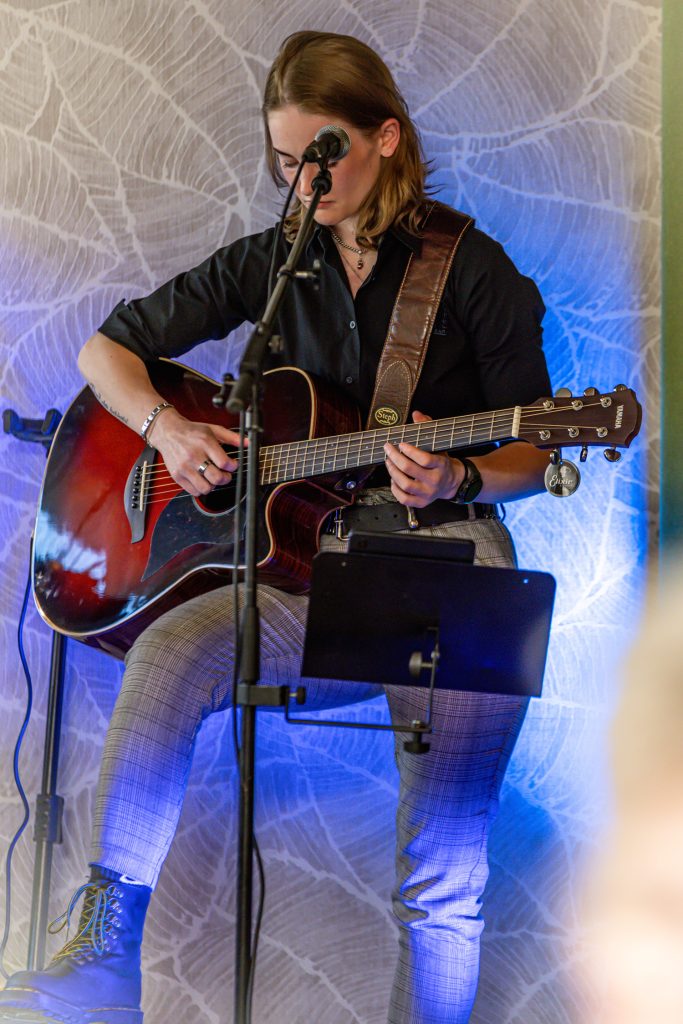 Eine Musikerin sitzt auf einem Hocker auf einer kleinen Bühne und spielt eine akustische Gitarre. Sie trägt ein schwarzes Hemd, karierte Hosen und dunkle Boots. Vor ihr steht ein Mikrofon, während blaues Licht die Hintergrundwand beleuchtet und eine intime Live-Musik-Atmosphäre schafft.