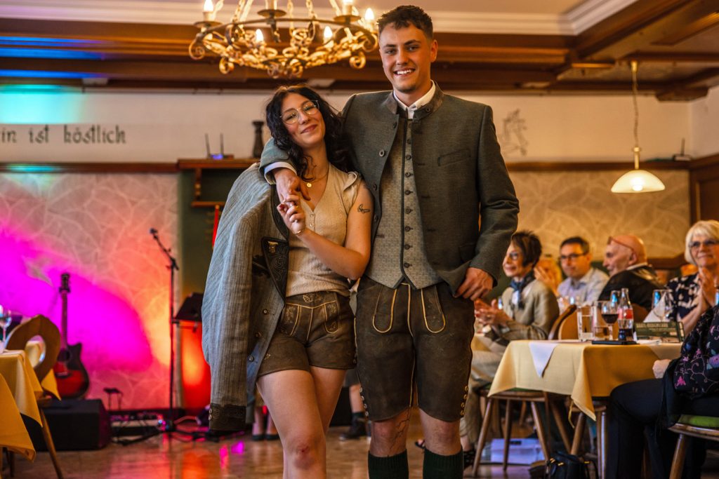 Ein junges Paar in Tracht steht bei einer Veranstaltung in einem Gastraum und lächelt in die Kamera. Beide tragen Lederhosen, der Mann zusätzlich eine Trachtenjacke. Gäste sitzen an den Tischen und beobachten die Szene. Im Hintergrund sind eine Bühne mit Beleuchtung und eine Gitarre zu sehen.