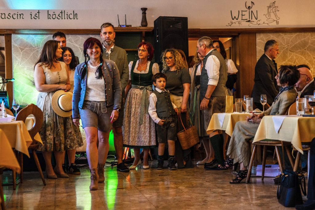 Mehrere Erwachsene und Kinder in Tracht stehen und gehen bei einer Veranstaltung durch einen Gastraum. Im Vordergrund läuft eine Frau in Lederhose und grauer Strickjacke lächelnd nach vorne. Gäste sitzen an den Tischen und beobachten die Szene. Der Raum ist mit Holzvertäfelungen, gedeckten Tischen und stimmungsvoller Beleuchtung gestaltet.