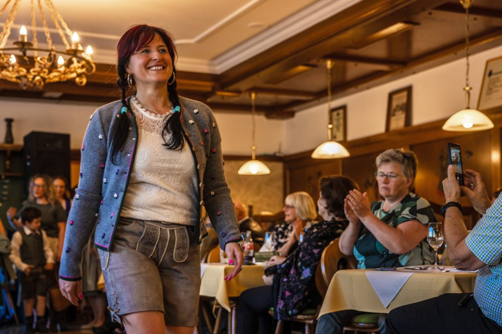 Eine Frau in Tracht mit Lederhose und grauer Strickjacke geht lächelnd bei einer Veranstaltung durch einen Gastraum. Gäste sitzen an den Tischen und applaudieren, eine Person fotografiert die Szene mit dem Smartphone. Im Hintergrund sind weitere Personen in Tracht zu sehen.