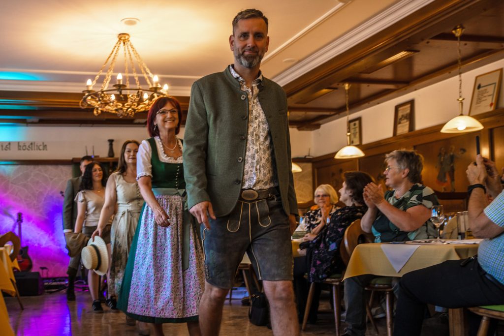 Ein Mann in Tracht mit grüner Jacke und Lederhose geht bei einer Veranstaltung durch einen Gastraum. Hinter ihm folgen weitere Personen in Tracht, darunter eine Frau in einem grünen Dirndl mit floral gemusterter Schürze. Gäste sitzen an den Tischen und applaudieren. Der Raum ist mit Holzvertäfelungen und Kronleuchtern gestaltet.