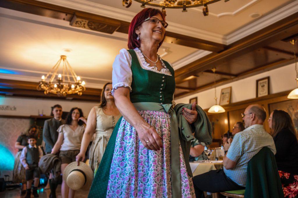 Eine Frau in einem grünen Dirndl mit floral gemusterter Schürze geht bei einer Veranstaltung durch einen Gastraum und lächelt. Hinter ihr folgen weitere Erwachsene und Kinder in Tracht. Gäste sitzen an den Tischen und beobachten die Szene. Der Raum ist mit Holzvertäfelungen und Kronleuchtern gestaltet.