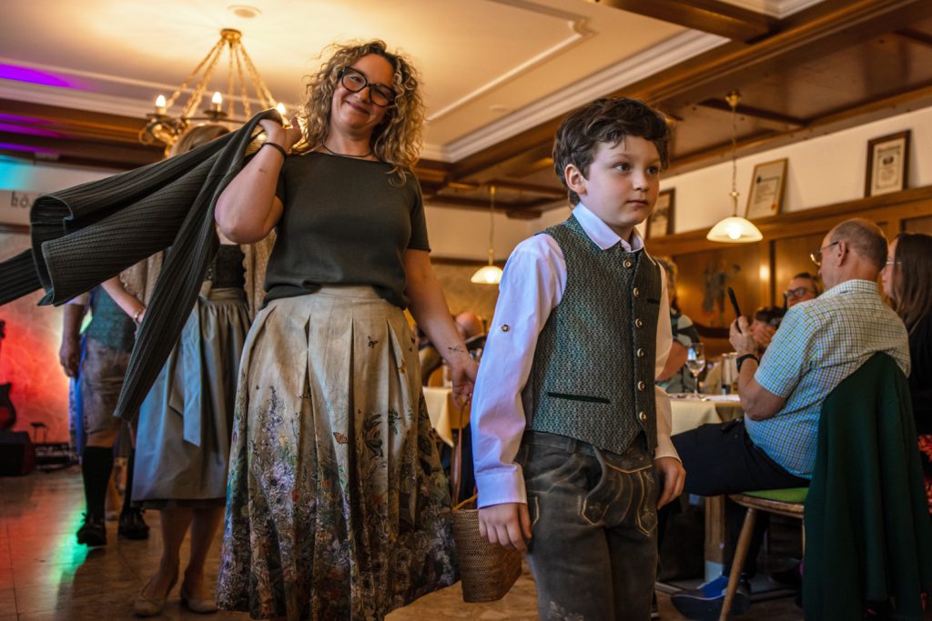 Eine Frau mit lockigen Haaren und ein Junge in Tracht gehen gemeinsam bei einer Veranstaltung durch einen Gastraum. Der Junge trägt Lederhose und Weste, die Frau hält eine Jacke über der Schulter. Im Hintergrund sind weitere Personen in Tracht zu sehen, Gäste sitzen an den Tischen und applaudieren.