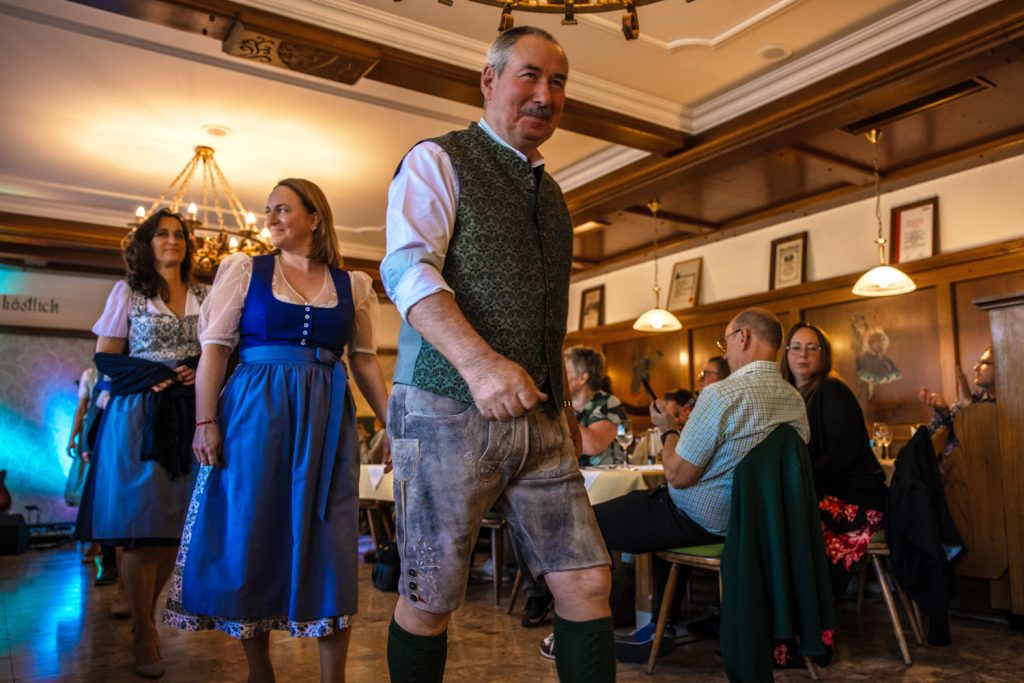 Ein Mann in Tracht mit Lederhose und Weste geht bei einer Veranstaltung durch einen Gastraum. Hinter ihm folgen zwei Frauen in Dirndln. Gäste sitzen an den Tischen und applaudieren. Der Raum ist mit Holzvertäfelungen und Kronleuchtern gestaltet.