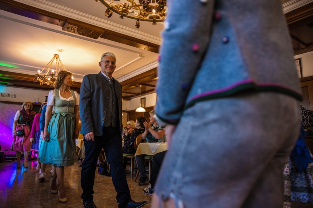 Mehrere Personen gehen bei einer Veranstaltung durch einen Gastraum, darunter eine Frau im türkisfarbenen Dirndl und ein Mann in Trachtenjacke. Gäste sitzen an den Tischen und applaudieren. Im Vordergrund ist unscharf eine weitere Person in Tracht zu sehen, der Raum ist mit Kronleuchtern und Holzvertäfelungen gestaltet.