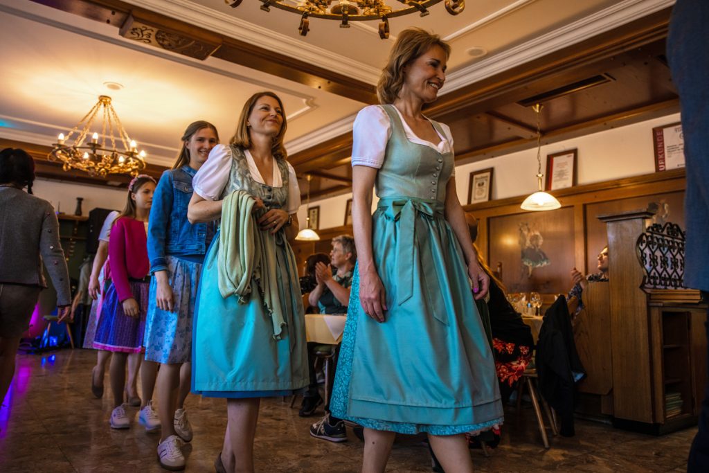 Mehrere Frauen und Mädchen gehen bei einer Veranstaltung durch einen Gastraum, einige tragen Dirndl in Türkis und Rosa. Gäste sitzen an den Tischen und applaudieren. Der Raum ist mit Holzvertäfelungen und Kronleuchtern ausgestattet.