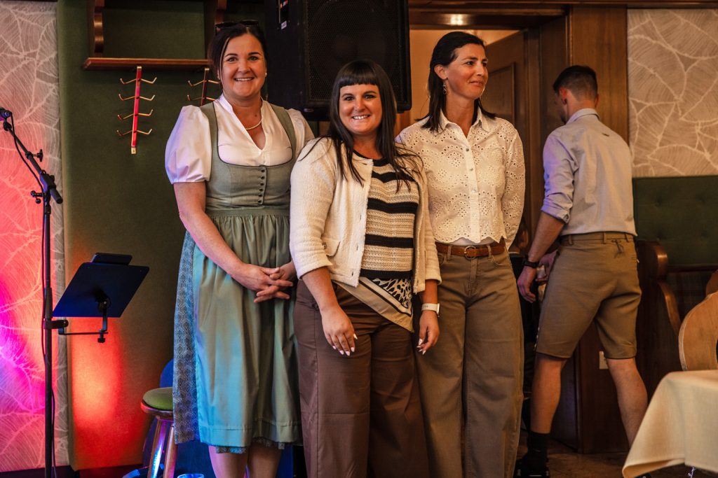 Drei Mitarbeiterinnen der Landmarkt Modehäuser stehen gemeinsam bei einer Veranstaltung und lächeln in die Kamera. Eine Frau trägt ein hellgrünes Dirndl, die anderen tragen Alltagskleidung. Im Hintergrund sind Lautsprecher und Bühnenbeleuchtung zu sehen, rechts geht ein Mann durch eine Tür.