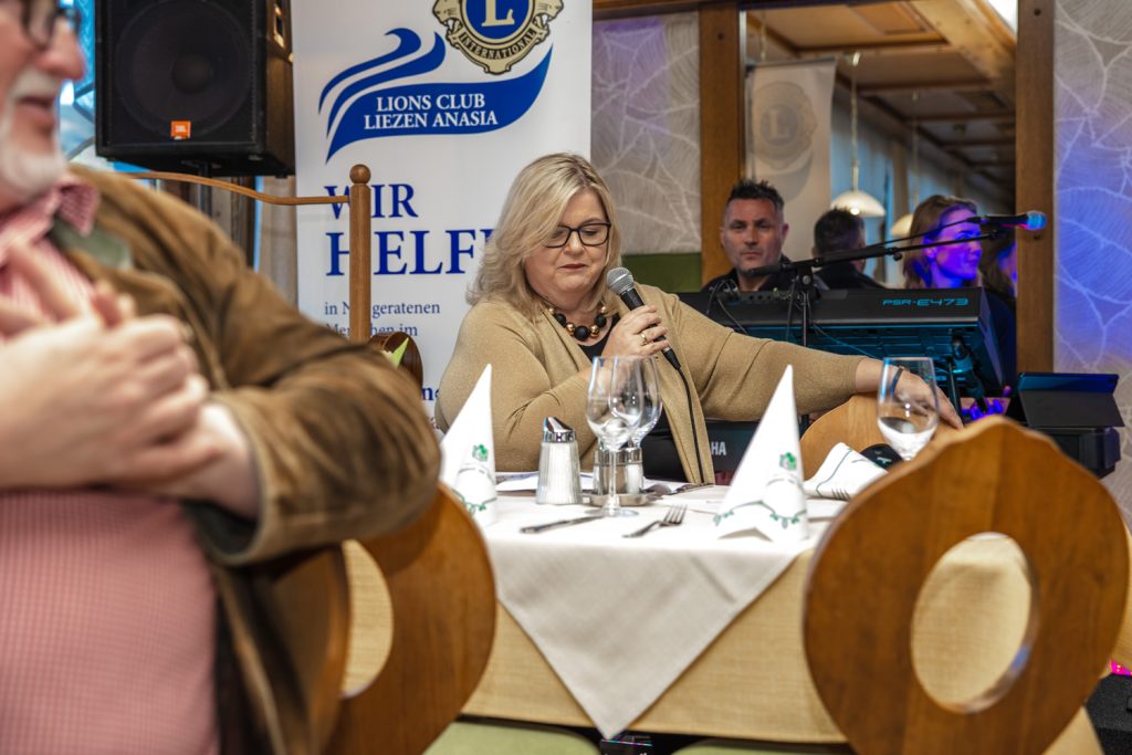 Eine Frau sitzt bei einer Veranstaltung des Lions Club Liezen Anasia an einem gedeckten Tisch und spricht in ein Mikrofon. Im Hintergrund sind Musiker mit Instrumenten sowie ein Banner des Lions Clubs zu sehen. Im Vordergrund sind unscharf zwei Personen und Holzstühle erkennbar.
