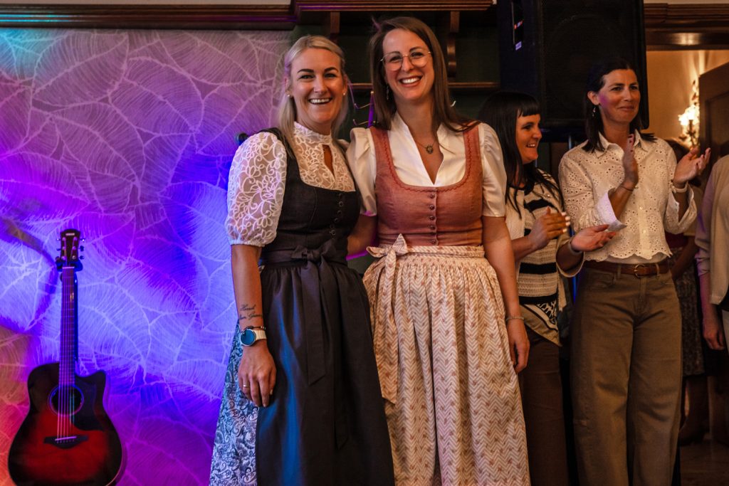 Mehrere Mitarbeiterinnen der Landmarkt Modehäuser stehen bei einer Veranstaltung gemeinsam vor einer beleuchteten Wand und lächeln in die Kamera. Zwei Frauen im Vordergrund tragen Dirndl in Rosa und Dunkelgrau. Im Hintergrund stehen weitere Frauen, einige applaudieren. Links im Bild ist eine Gitarre zu sehen.