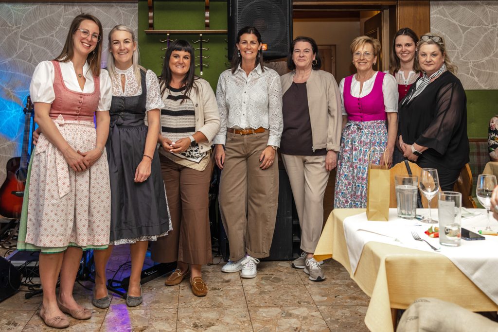 Mehrere Mitarbeiterinnen der Landmarkt Modehäuser stehen gemeinsam bei einer Veranstaltung in einem Gastraum und lächeln in die Kamera. Einige tragen Dirndl, andere Alltagskleidung. Im Vordergrund sind gedeckte Tische mit Gläsern und Servietten zu sehen, im Hintergrund Lautsprecher und Musikinstrumente.
