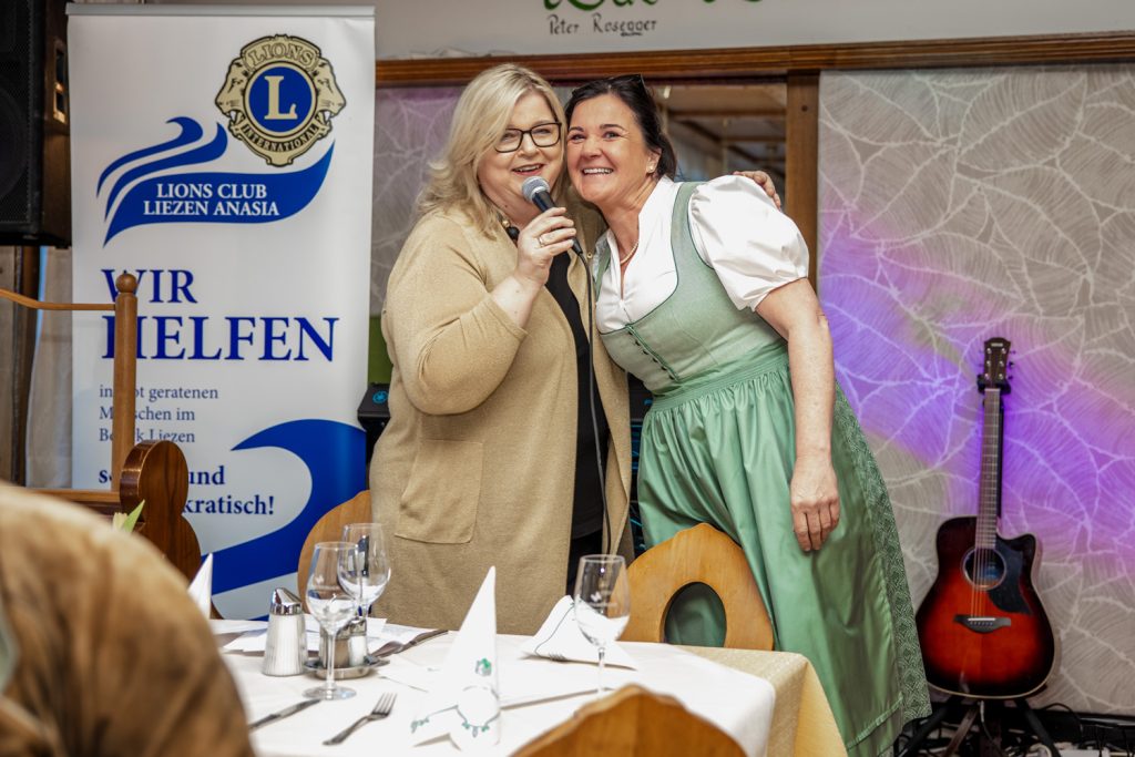 Zwei Frauen stehen eng beieinander bei einer Veranstaltung des Lions Club Liezen Anasia. Eine Frau spricht lächelnd in ein Mikrofon, die andere trägt ein hellgrünes Dirndl und lehnt sich an sie. Im Hintergrund sind ein Banner des Lions Clubs und eine Gitarre zu sehen, im Vordergrund gedeckte Tische mit Gläsern und Servietten.