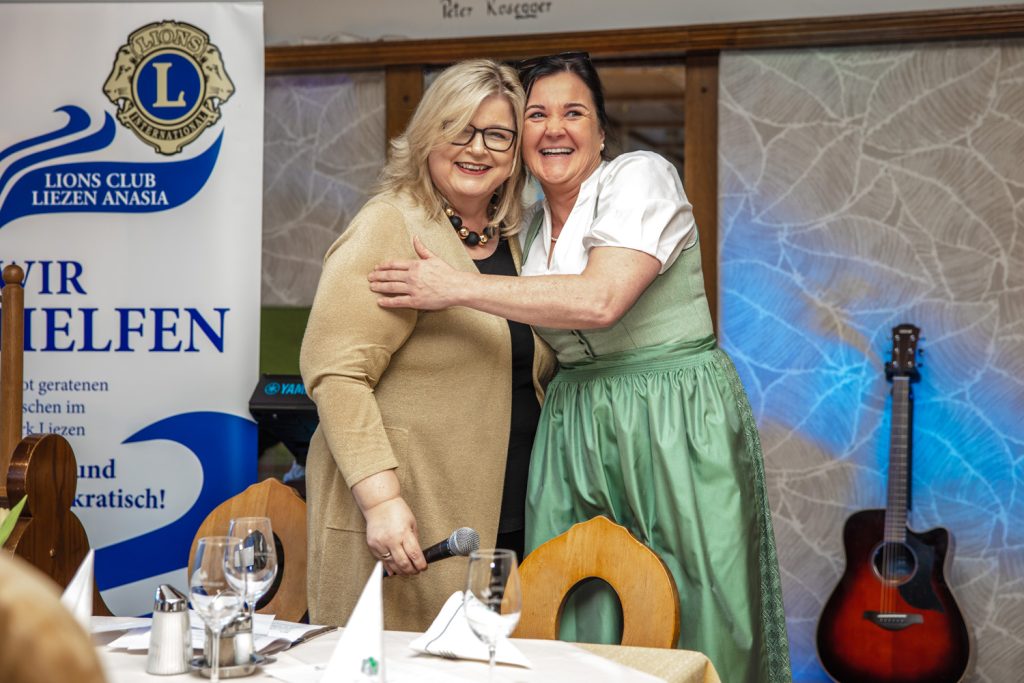 Zwei Frauen umarmen sich lachend bei einer Veranstaltung des Lions Club Liezen Anasia. Eine Frau hält ein Mikrofon in der Hand, die andere trägt ein hellgrünes Dirndl. Im Hintergrund stehen ein Banner des Lions Clubs und eine Gitarre, im Vordergrund sind gedeckte Tische mit Gläsern und Servietten zu sehen.