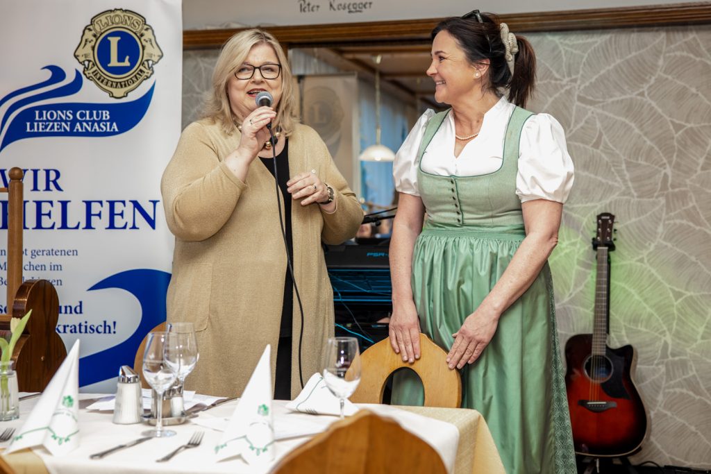 Zwei Frauen stehen bei einer Veranstaltung des Lions Club Liezen Anasia in einem Gastraum. Eine Frau spricht mit einem Mikrofon, die andere trägt ein hellgrünes Dirndl und lächelt sie an. Im Hintergrund sind ein Banner des Lions Clubs sowie eine Gitarre zu sehen. Im Vordergrund stehen gedeckte Tische mit Gläsern und Servietten.