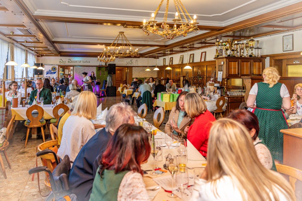 Gemütliche Veranstaltung in einem großen, holzvertäfelten Saal mit Kronleuchtern und festlich gedeckten Tischen. Zahlreiche Gäste sitzen zusammen, unterhalten sich und genießen die Atmosphäre, während im Hintergrund eine kleine Bühne mit Musik und Technik aufgebaut ist.