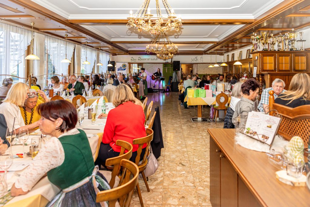Großer Veranstaltungssaal mit zahlreichen Gästen an festlich gedeckten Tischen während eines Events des Lions Club Liezen Anasia. Im Hintergrund sorgt Live-Musik für Stimmung, während sich die Besucher unterhalten und das gemeinsame Beisammensein genießen.
